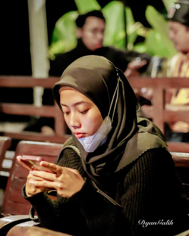 A young woman wearing a hijab is playing with her gadget against blur background. Gear: Canon EOS Rp. Lens: Canon 50mm. Model: -. Spot: Warung Kulinan. #candid #portrait #closeup. Owner: @DyanGalih